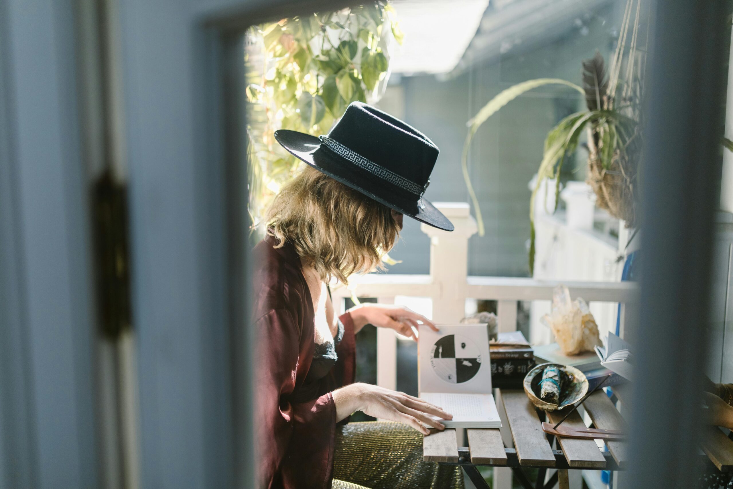 A woman in a hat reading tarot cards on a sunlit balcony surrounded by crystals.